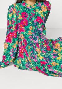 Blumenkleid mit grünem Hintergrund, mit rosa, gelben und lila Blumen, Knopfverschluss und langen Ärmeln. Weicher, fließender Stoff.