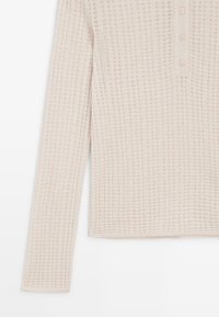 Jasnobeżowa bluzka z długim rękawem o fakturze przypominającej wafle, z zapięciem na guziki przy dekolcie i prostymi rękawami.