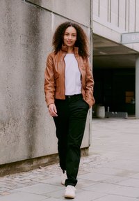 Woman with curly hair wearing a brown jacket, white shirt, black pants, and white sneakers walking on an urban sidewalk near a concrete wall.