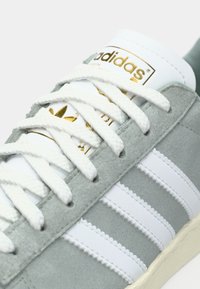 Grijze suède sneaker met witte leren strepen, witte veters en een goudkleurig logo op de tong. Gestructureerd oppervlak en klassiek ontwerp.