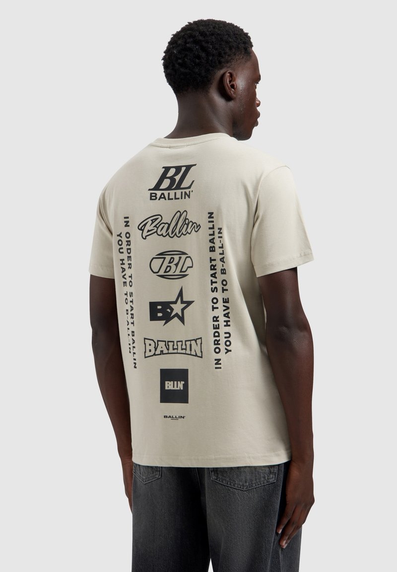 Man die van achteren te zien is, draagt een beige T-shirt met meerdere zwarte "Ballin'" logo's en tekst op de achterkant, gecombineerd met een donkere spijkerbroek.