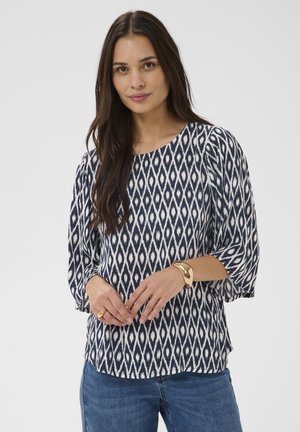 Kobieta o długich ciemnych włosach, ubrana w bluzkę z czarno-białym wzorem w romby, niebieskie dżinsy oraz złotą bransoletkę i pierścionki, stojąca na białym tle.