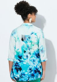 Blusa floreale in azzurro chiaro e bianco, caratterizzata da grandi fiori blu e un design con schiena plissè. Maniche lunghe arrotolate ai polsi.