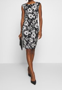 Czarna sukienka w kwiaty z białymi kwiatami, bez rękawów, dopasowana, długości do kolan, w połączeniu z czarną kopertówką i wysokimi obcasami.