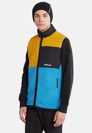 Gilet à blocs de couleurs avec du polaire jaune, du polaire bleu et une poche en nylon noir. Fermeture éclair, poches latérales et col haut.
