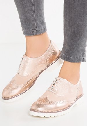 Metallic roseguld snørebåndssko med brogue-detaljer, tekstureret læder og en hvid gummisål. Bruges med grå opfoldede bukser.