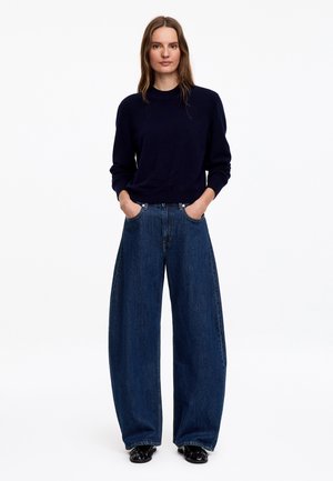 Femme portant un haut noir à manches longues et un jean bleu à jambes larges, les mains dans les poches, debout devant un fond blanc uni.