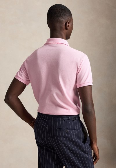 Uomo che indossa una polo rosa chiaro a maniche corte e pantaloni blu scuro a righe sottili, in piedi con una mano sul fianco.