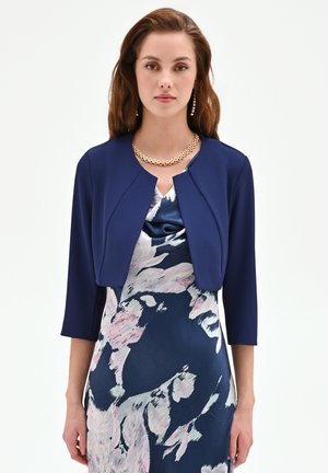 Giovane donna che indossa una giacca corta blu navy sopra un vestito floreale blu navy e rosa, accessoriata con una collana a catena dorata e orecchini a goccia.