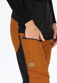 Pantaloni arancioni e neri con tasche con zip, una finitura testurizzata e una toppa rettangolare con logo sulla coscia, progettati per l'uso all'aperto.