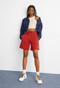 Shorts en coton rouges avec une taille élastique et des poches latérales, associés à un débardeur court beige et une veste en jean bleue. Baskets blanches.