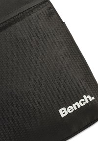 Schwarzer Stoff mit sechseckigem Muster, ausgestattet mit einer Reißverschlusstasche und einem weißen "Bench."-Logo in der unteren rechten Ecke. Strukturiertes Finish.
