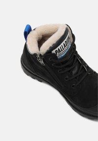 Bota de tornozelo em camurça preta com um interior peludo. Apresenta um fecho lateral com zíper e atacadores, etiqueta da marca e uma alça azul no calcanhar.