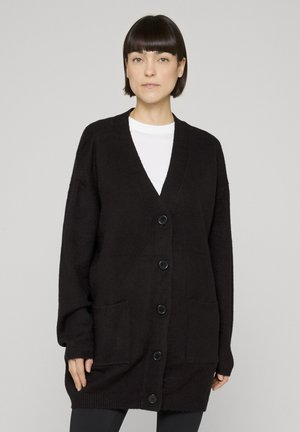 Donna con capelli corti e scuri che indossa una maglietta bianca sotto un maxi cardigan nero con bottoni e tasche frontali, in piedi davanti a uno sfondo chiaro e uniforme.