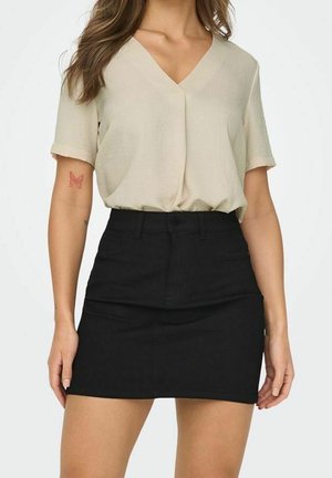 Kobieta ubrana w beżową bluzkę z krótkim rękawem i dekoltem w serek, wpuszczoną w czarną mini spódnicę z wysokim stanem, stoi z rękami swobodnie opuszczonymi wzdłuż boków ciała.