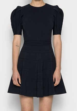 Woman wearing a short black dress with puffed short sleeves and horizontal seam details on the skirt and waist.
