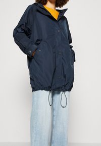 Personne portant une veste à capuche oversize bleu marine avec des boutons sur le devant, des cordons et des poches, associée à un jean en denim large bleu clair et un pull jaune.