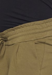Pantalons vert olive en tissu doux et texturé. Dotés d'une taille à cordon de serrage et de plis à l'avant. Associés à une chemise noire au-dessus.