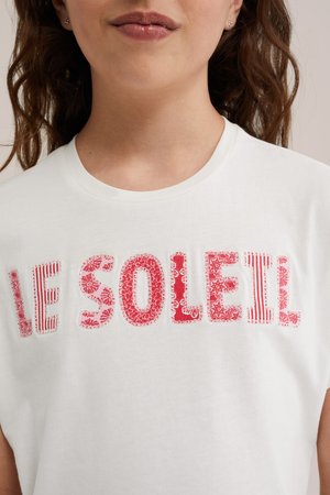 Donna con camicia bianca con la scritta "LE SOLEIL" in lettere rosse decorate sul petto, mostrando labbra, collo e parte superiore del busto.