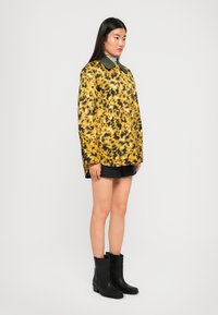 Cappotto a stampa leopardata gialla con colletto verde, maniche lunghe e due tasche frontali, abbinato a una mini gonna nera e stivaletti neri.
