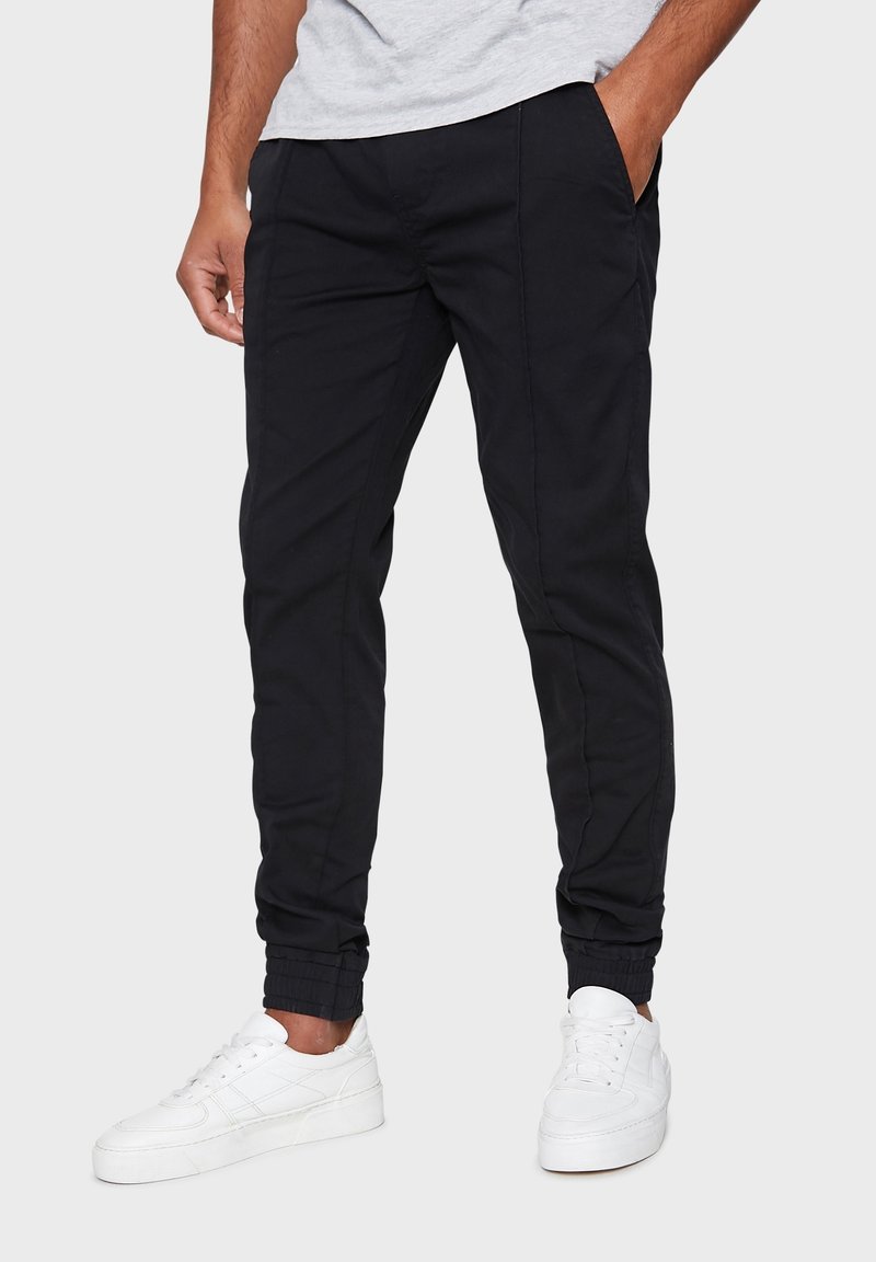 Pantaloni jogger neri con una vestibilità slim, dotati di tasche laterali e polsini elasticizzati, abbinati a sneaker bianche e una maglietta grigia chiara.