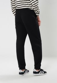 Sorte joggebukser med elastiske sluttninger, med en jevn tekstur. Båret med svart og hvitstripete langermet skjorte og svarte joggesko.