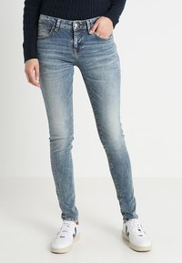 Slim-fit ljusblå jeans som bärs med vita sneakers och en mörk marinblå grovstickad tröja, person som står mot en enfärgad bakgrund.