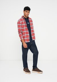 Camicia a quadri rossa e bianca sopra una t-shirt blu navy, abbinata a jeans in denim scuro e sneakers marroni con dettagli bianchi.