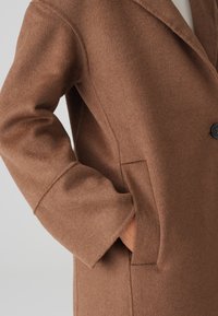 Brown wool blend coat with a notched collar, large side pocket, and a single black button closure. Soft texture and relaxed fit.
