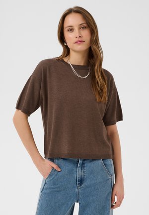 Kvinde med langt brunt hår iført en løs brun kortærmet sweater og lyseblå jeans, stående med den ene hånd i lommen mod hvid baggrund.