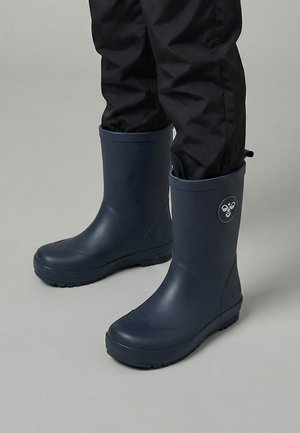 Navyblauwe rubberen regenlaarzen gedragen met zwarte broek, met een rond bijenlogo aan de buitenkant van elke laars.