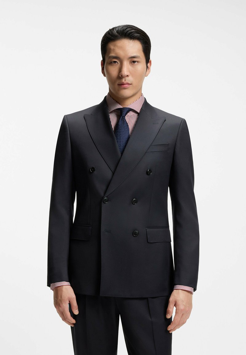Homme portant un costume croisé sombre avec une chemise rayée et une cravate texturée foncée, debout devant un fond blanc uni.