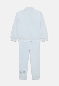 Survêtement en velours bleu clair comprenant une veste à col et un pantalon assorti. Le pantalon est brodé de "ESS" sur la jambe gauche. Texture douce.