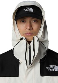 White and black jacket with hood, featuring zipped front, high collar, and logo accents. The fabric appears durable with a smooth texture.