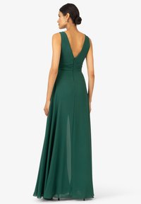 Langes grünes Abendkleid aus fließendem Stoff, mit V-Ausschnitt im Rücken, breiten Trägern, geraffter Taille und einem seitlichen Schlitz für Bewegungsfreiheit und Eleganz.
