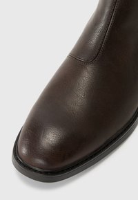 Botín de cuero marrón con textura suave, punta redondeada y costuras contrastantes a lo largo de la costura, con suela de goma negra.