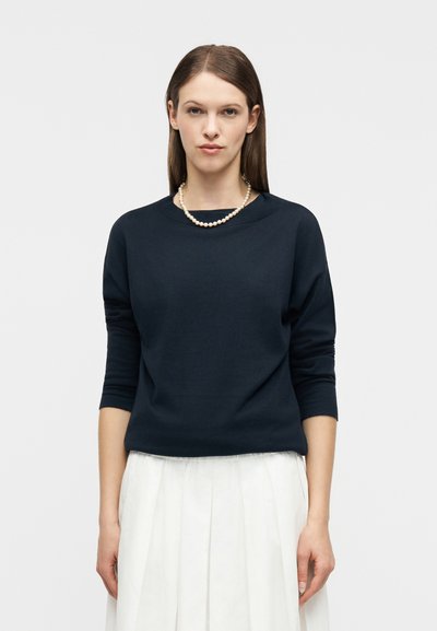 Femme aux cheveux bruns raides portant un haut noir à manches longues, une jupe blanche plissée et un collier de perles, debout devant un fond blanc uni.
