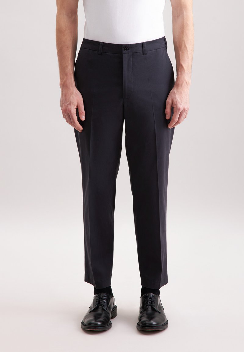 Ciemnonawowe spodnie charakteryzują się dopasowanym krojem, gładką fakturą, pętelkami na pasek oraz zapięciem na jeden guzik, w połączeniu z czarnymi butami.