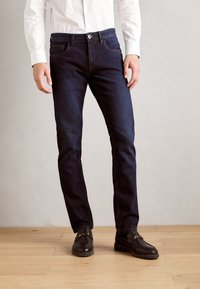 Jeans in denim blu scuro con design a gamba dritta, cinque tasche e cuciture a contrasto. Abbinati a mocassini marroni e una camicia bianca.