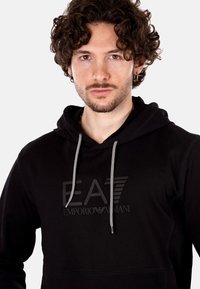 Felpa nera in cotone con tasca a marsupio, cappuccio con cordino e logo embossato "EA7 Emporio Armani" sul davanti.