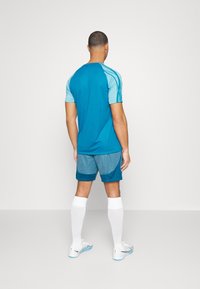 Hombre de pie de espaldas, vistiendo una camisa de manga corta azul y celeste, pantalones cortos azules, calcetines blancos hasta la rodilla y zapatillas blancas.
