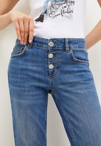 Mujer que lleva jeans de talle alto azules con tres botones decorativos plateados y una camiseta blanca con gráfico que muestra texto y una pierna ilustrada.