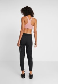 Czarne sportowe spodnie z odblaskowymi zamkami błyskawicznymi, zestawione z jasnoróżowym sportowym biustonoszem z wycięciem na plecach. Strój podkreśla dopasowany krój i gładką teksturę.