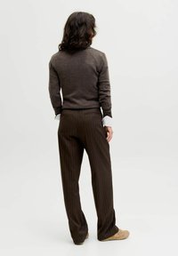 Osoba s tamnosmešnom kovrdžavom kosom stoji okrenuta leđima, obučena u smeđi pulover, bijele rukave košulje, smeđe hlače na tanke pruge i bež cipele.