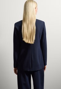 Blazer navy su misura con spalline, apertura posteriore e polsini con bottoni, abbinato a pantaloni coordinati. Texture liscia e design strutturato.