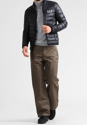 Man draagt een zwarte doorgestikte jas over een grijze coltrui, bruine wijde broekspijpen en zwarte sneakers, staand tegen een effen achtergrond.