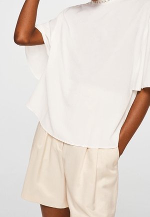 Top bianco a maniche corte con un design fluido, indossato con pantaloni corti plissettati beige. Tessuto morbido, vestibilità comoda e superficie liscia.