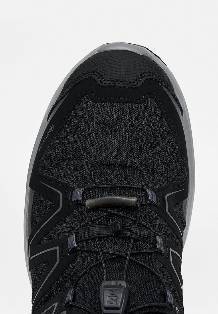 Zapatilla deportiva negra con suela gris, parte superior de tela de malla, sistema de cordones de ajuste rápido y etiqueta Gore-Tex sobre fondo claro.