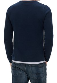 Mies, joka on pukeutunut tummansiniseen muotoiltuun pitkähihaiseen neuleeseen ja tummansiinisiin farkkuihin, käsivarret taskuissa, kuvattu takaa.