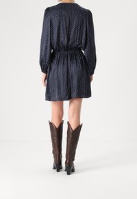 Nainen, jolla on yllään tummansininen pitkähihainen mini-mekko, jossa on pieniä pilkkuja, ja tummanruskeat polven yli ulottuvat korkeat saappaat, seisoo valkoisella lattialla.